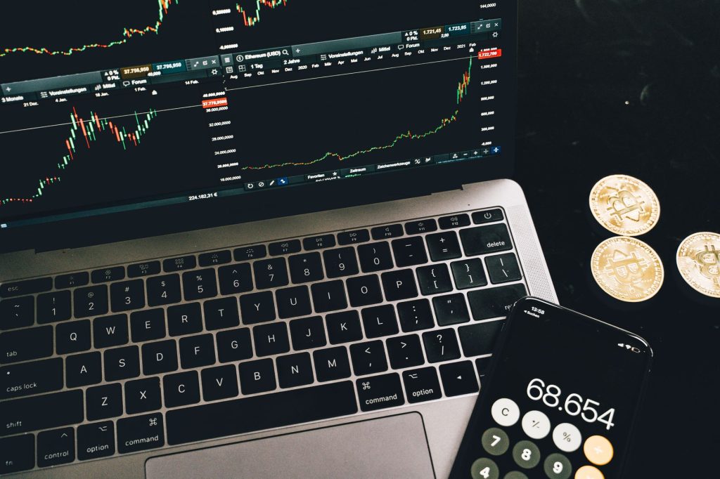 A laptop with trading charts, smartphone calculator, and bitcoin coins depicting cryptocurrency trading.